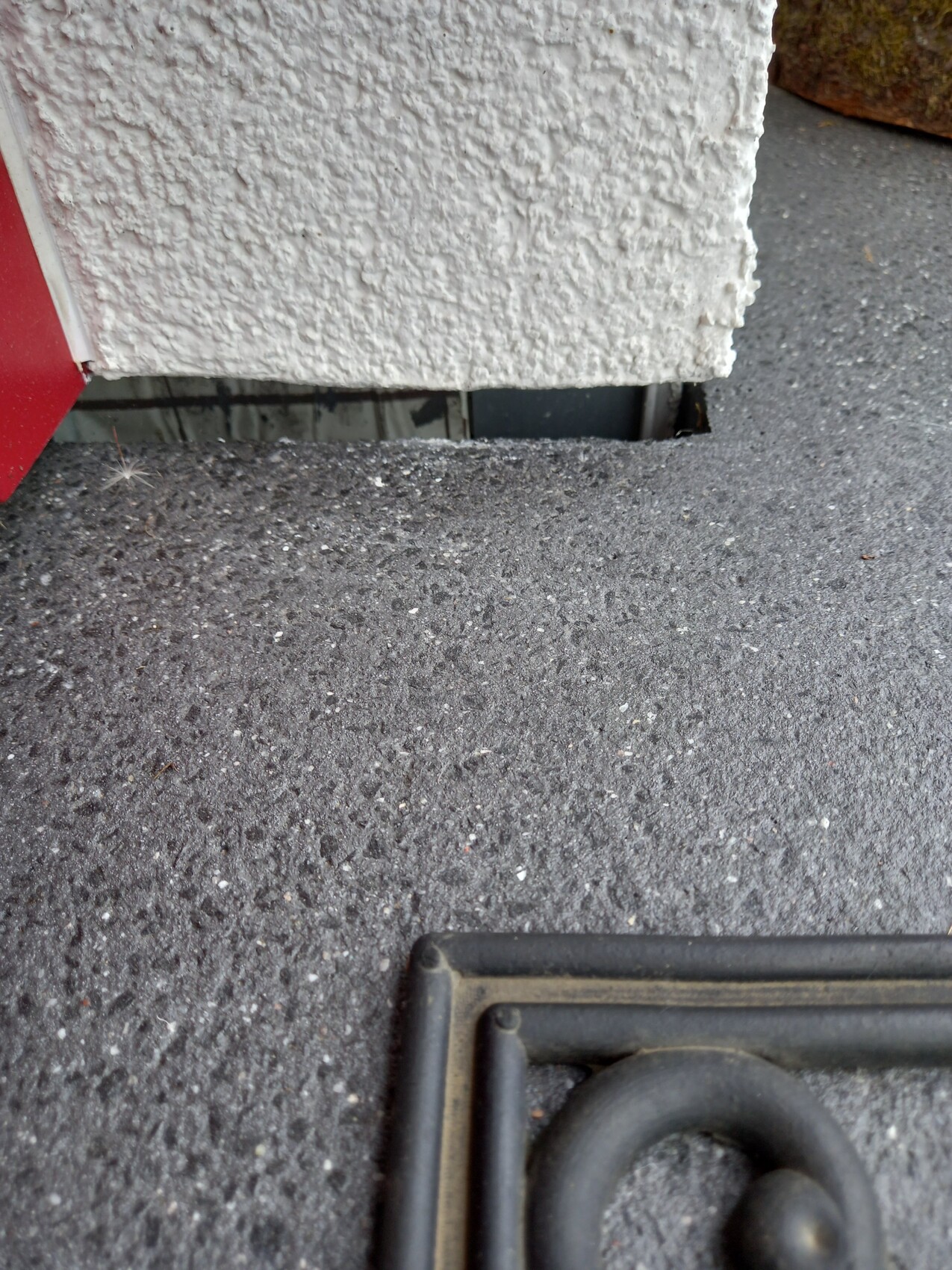 Außenansicht: weiße raue Wand, darunter Spalte; grauer Boden; roter Rand links; runder Metallring rechts