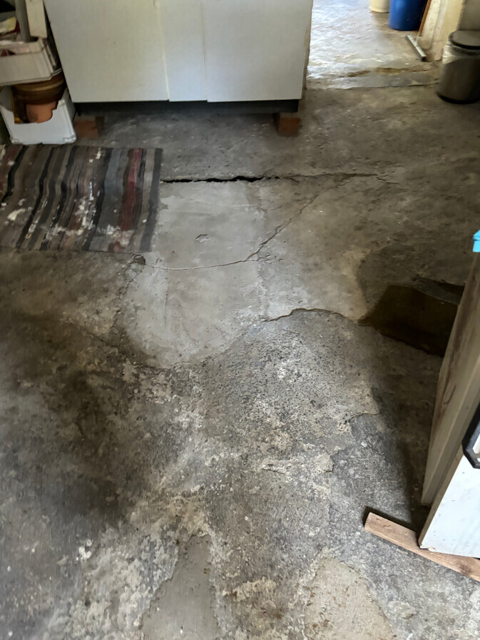 Verschmutzter Betonboden mit Rissen in einem Keller; links Teppich, hinten weißer Schrank.