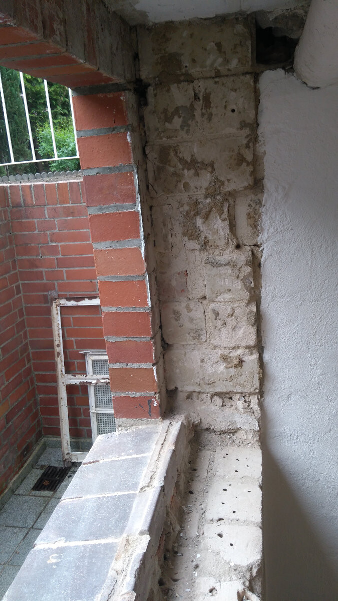 Außenwand aus unregelmäßigen Steinblöcken neben rotem Ziegelbau, Balkonbrüstung sichtbar.