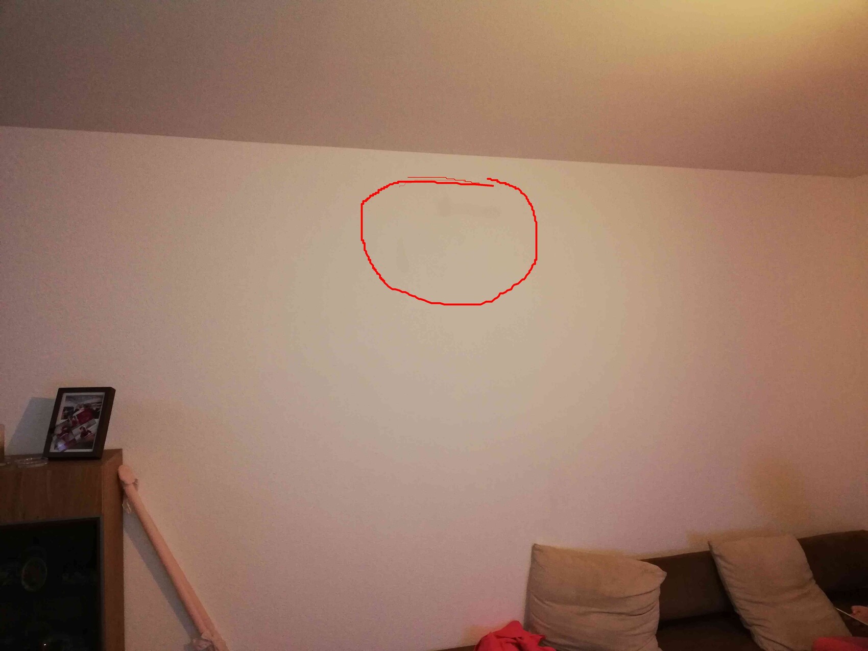 Wohnzimmer: Couch rechts, Schrank links mit Foto; roter Kreis markiert Wandfleck in Mitte.