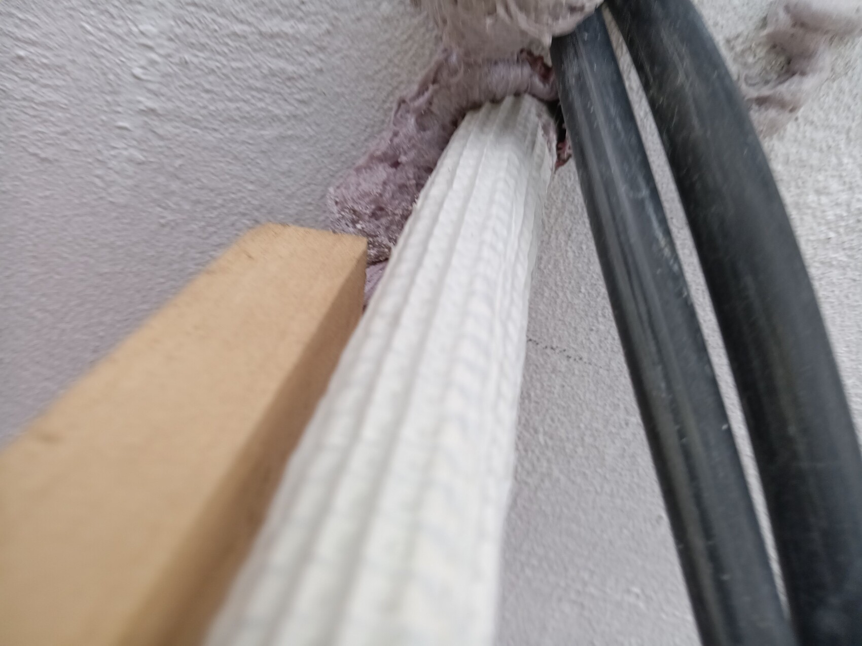 Nahaufnahme einer Wand mit schwarzem Kabel und einer weißen, geriffelten Leiste neben rohem Putz.