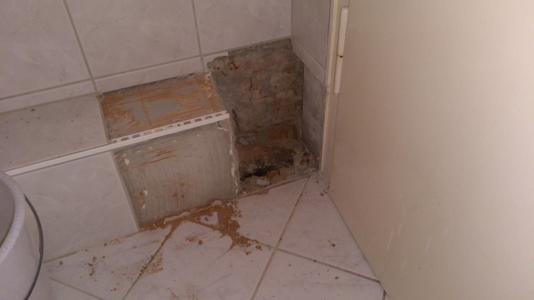 Badezimmer-Ecke mit freigelegter Wand aus Ziegel/Beton, Schutt auf Fliesen, Tür rechts.