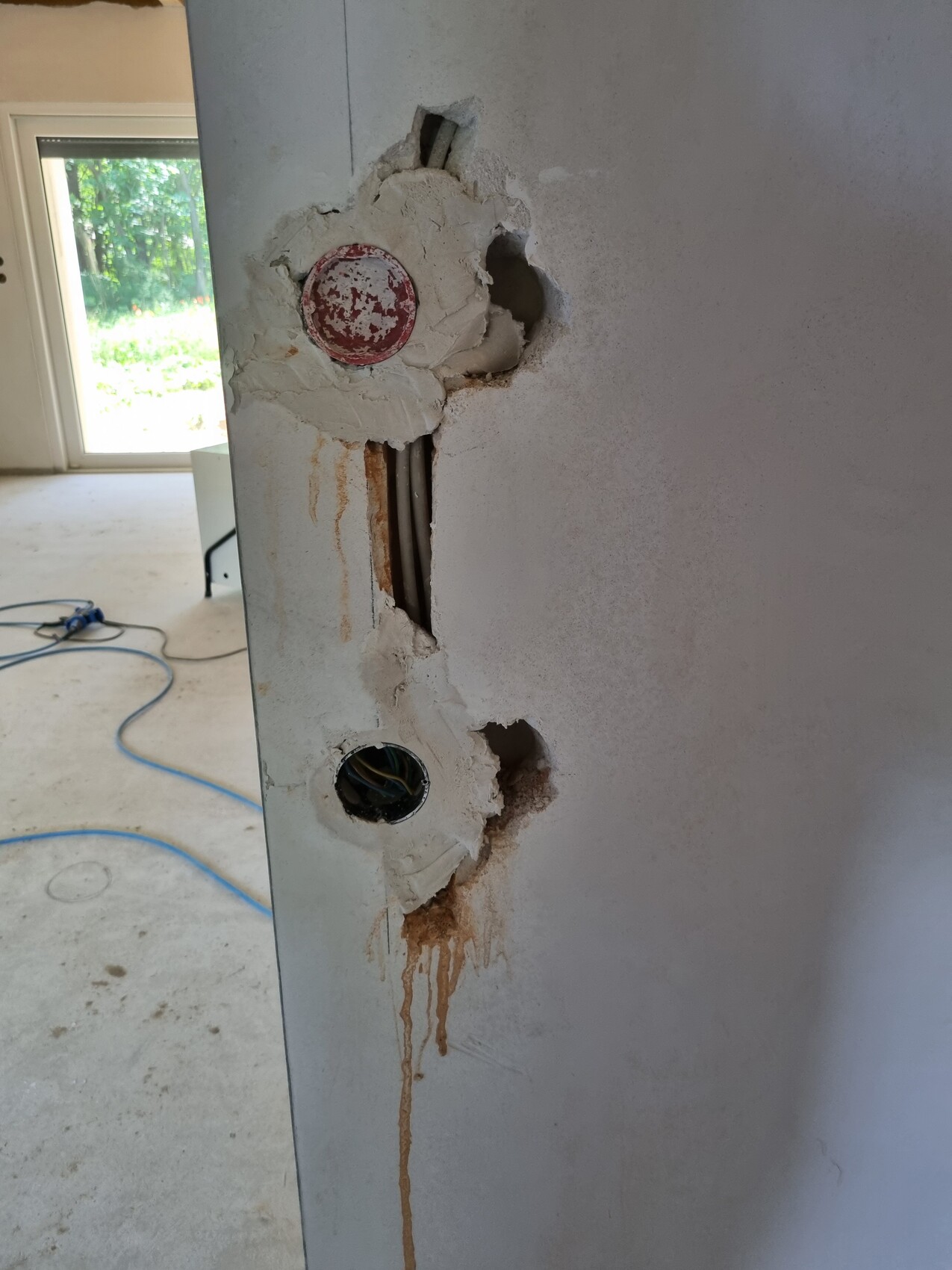 Wand mit Löchern und Putzrissen, rote Abdeckkappe, freiliegte Rohre, Rostflecken und Kabel.