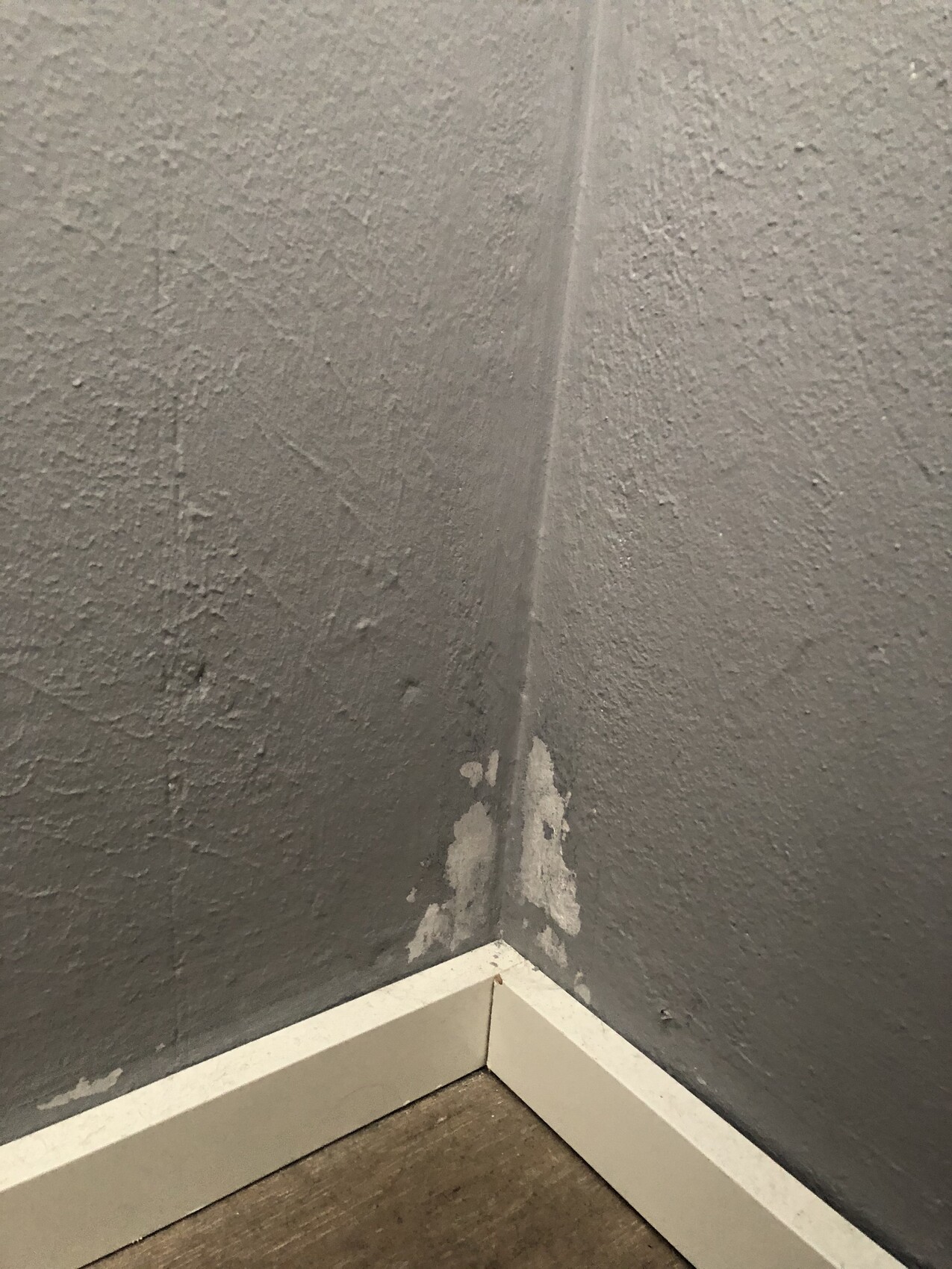 Ecke zweier grauer Wände mit abgeplatzter Farbe/Verputz nahe der weißen Sockelleiste.