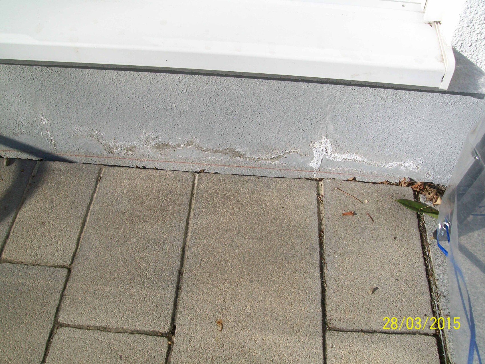 Grauer Pflasterboden vor rauer weißer Wand mit Rissen und Ablagerungen, Blattreste.