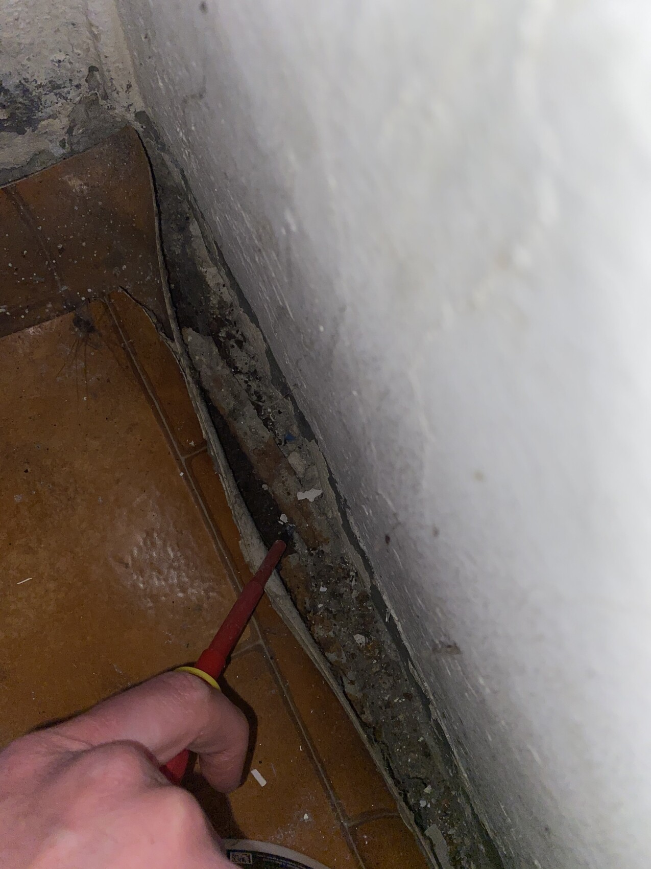 Nahaufnahme einer Hand mit rotem Schraubendreher an der staubigen Boden-Wand-Fuge zwischen Fliesen.