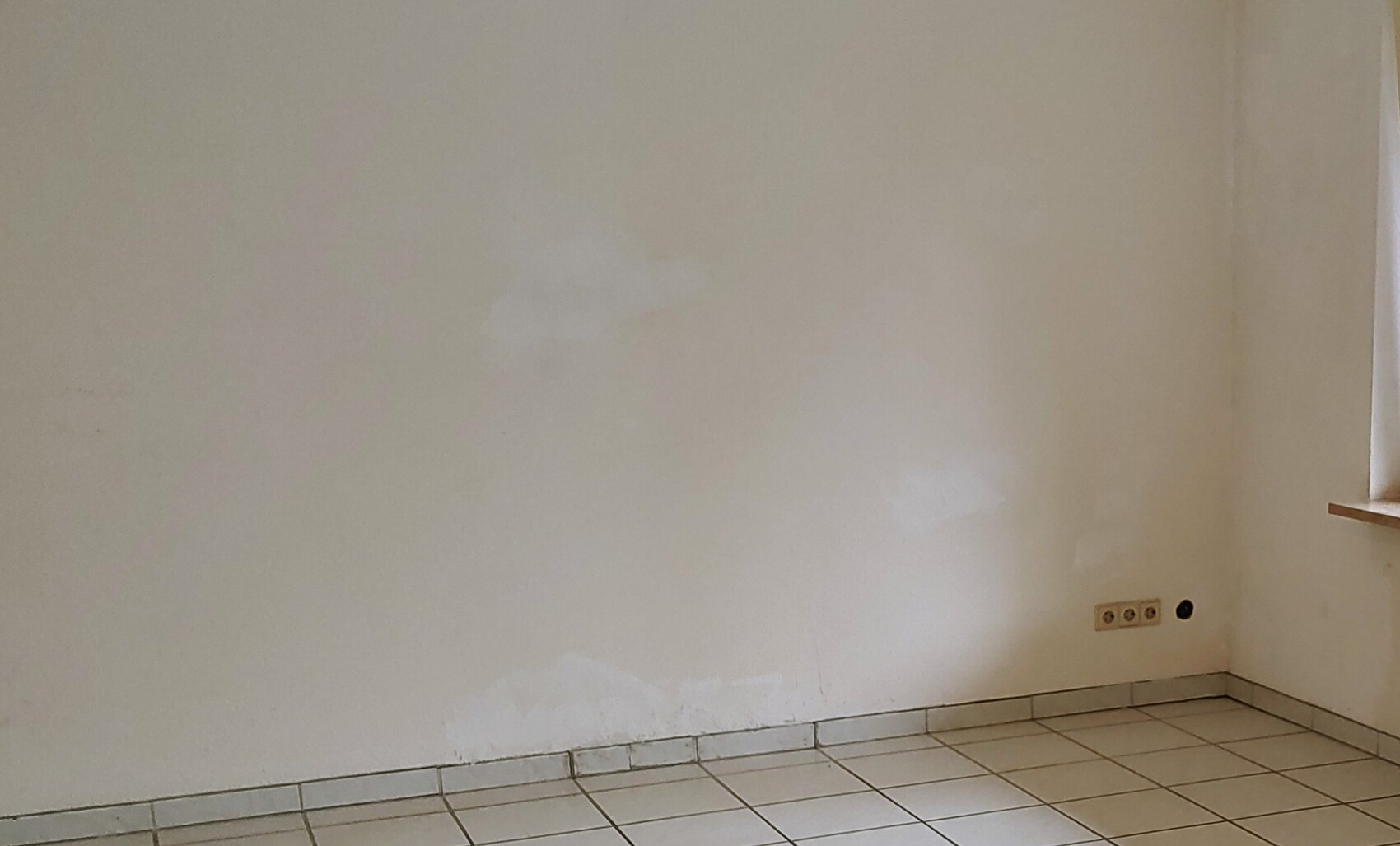Leerer Innenraum mit weißen Wänden und Putzresten, Fliesenboden, Steckdosen rechts unten.