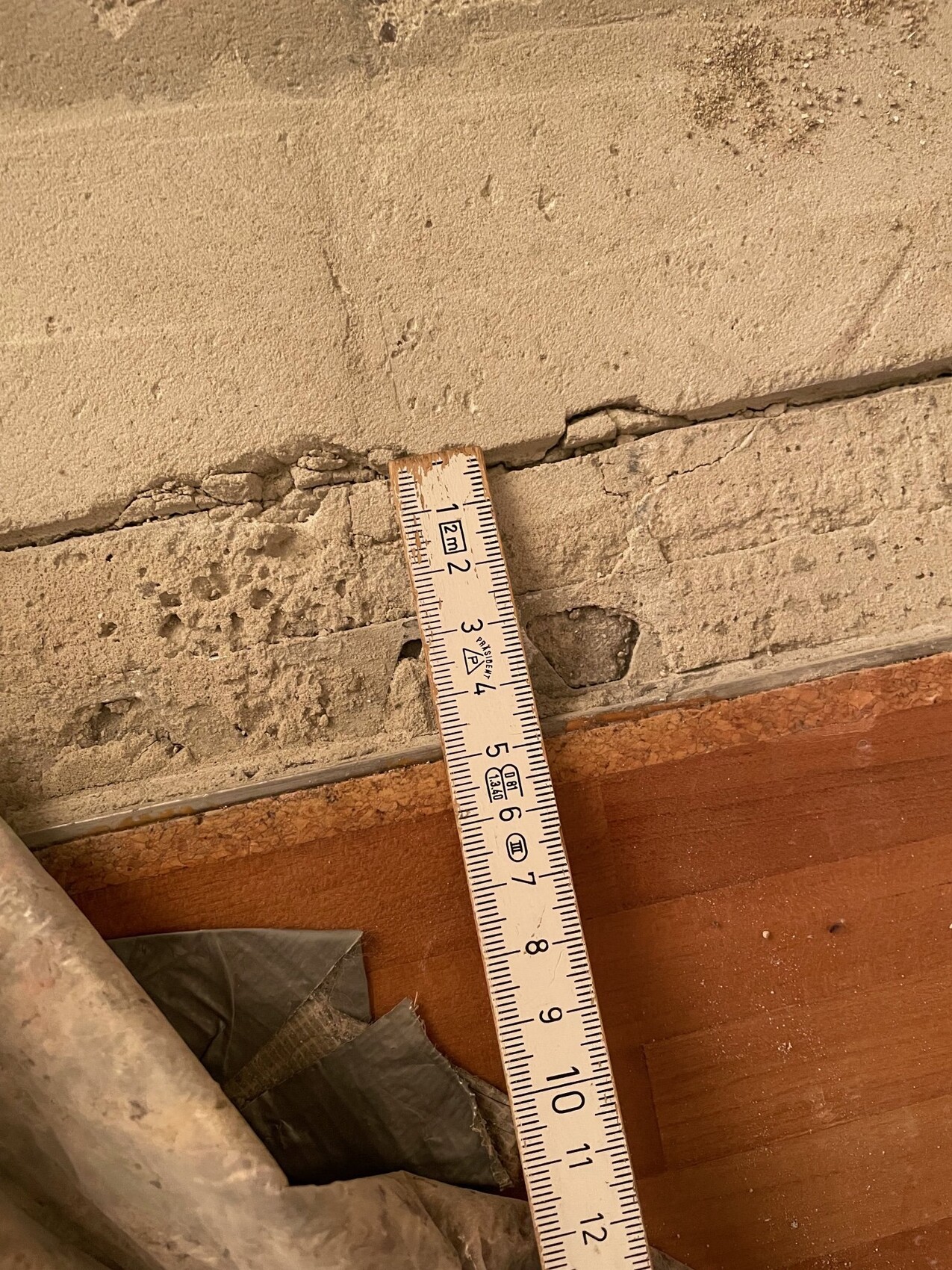Schräg stehendes Maßband an einer rissigen, verputzten Wand über einem Holzboden.