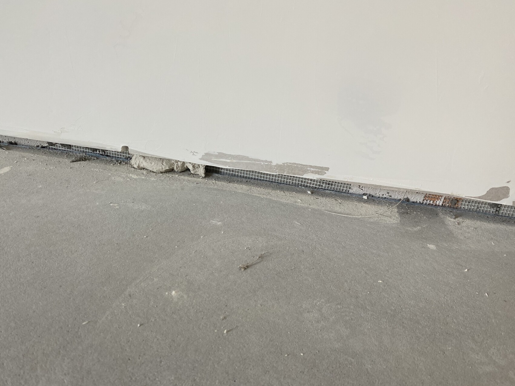 Weiße Wand mit abplatzendem Putz, bodennah Metallgitterleiste und Schutt am Boden.
