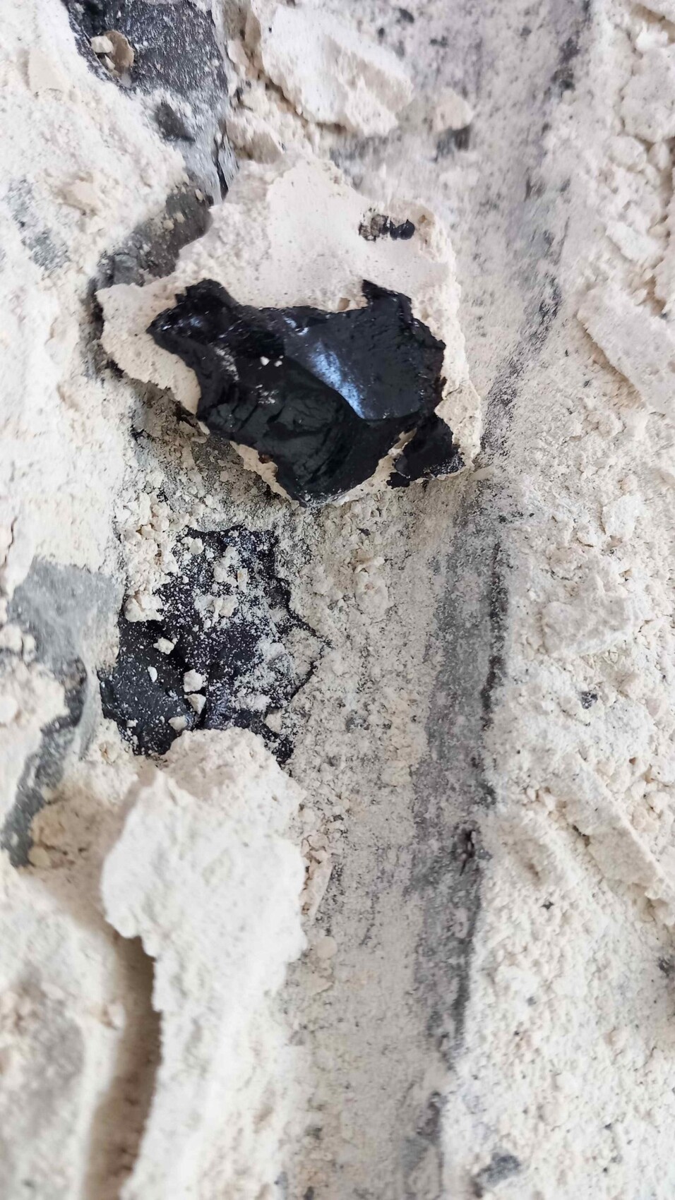 Nahaufnahme eines schwarzen, glänzenden Gesteinsstücks, umgeben von hellbeigem, bröckeligem Staub.