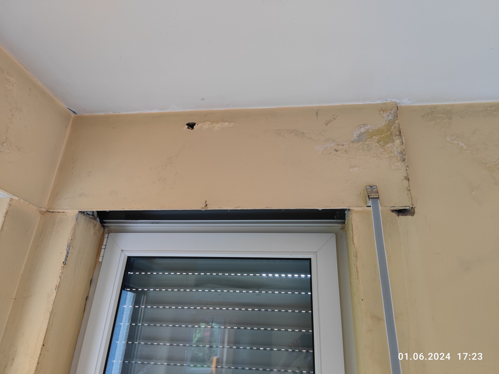 Fenster mit weißem Rahmen an gelber Wand; Risse, Feuchtigkeit, Loch oben, Kabelkanal rechts.