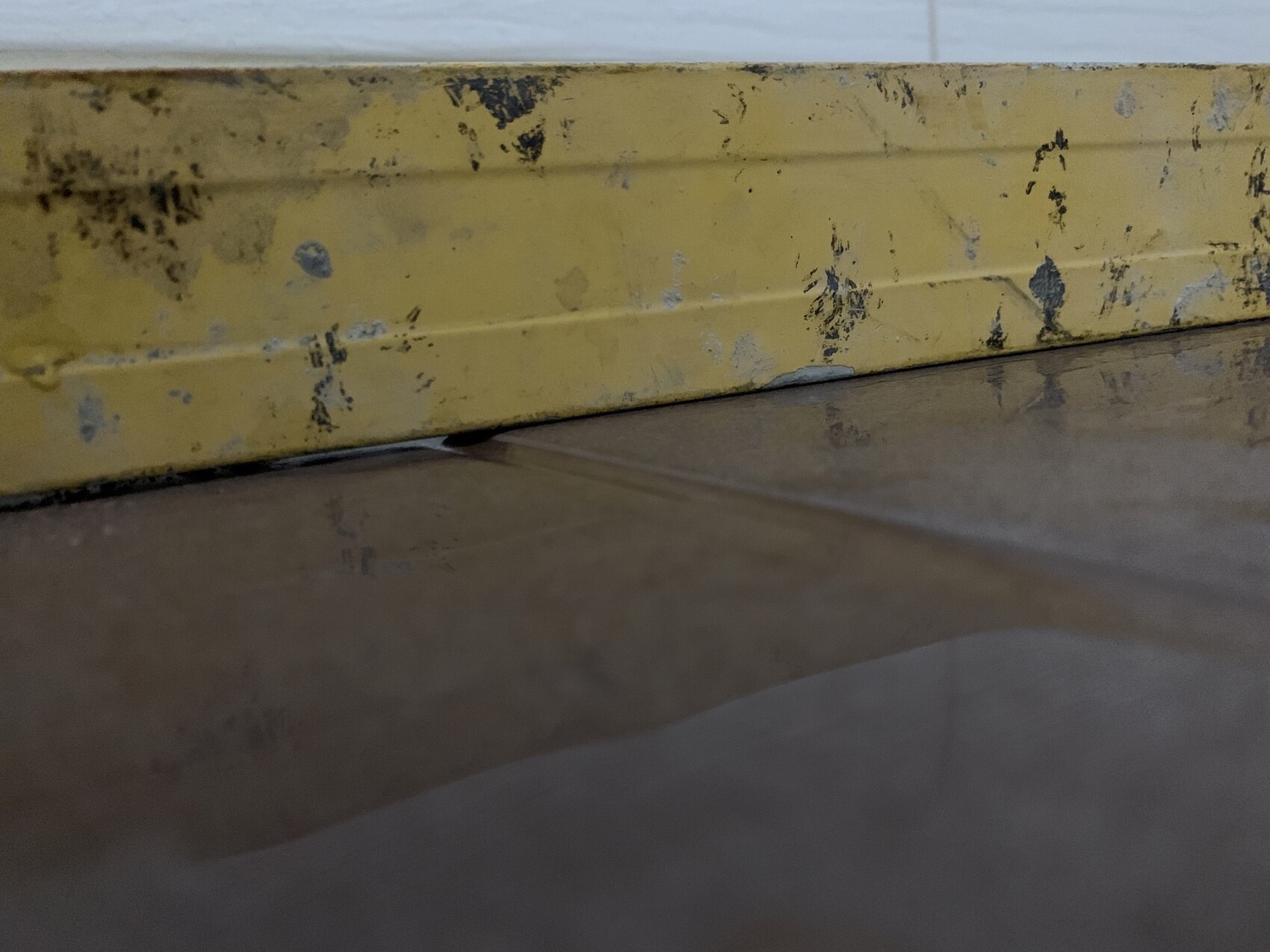Gelbe Sockelleiste mit schwarzen Flecken an einer Wand; darunter feuchter, reflektierender Boden.