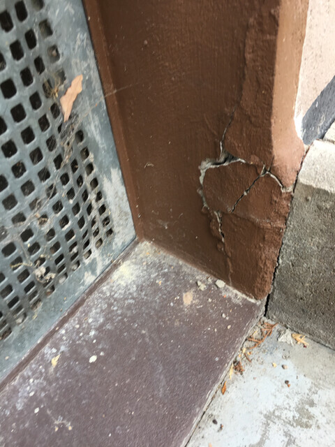 Außenwand Ecke mit braunem Putz und Riss neben Metallgitter.