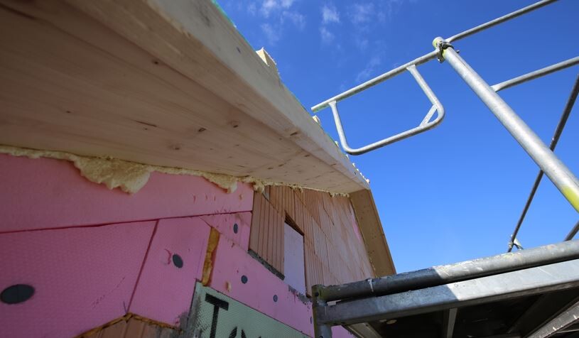Baustelle: Pinke Dämmplatten an Wand, Holzverkleidung, Gerüst gegen blauen Himmel.