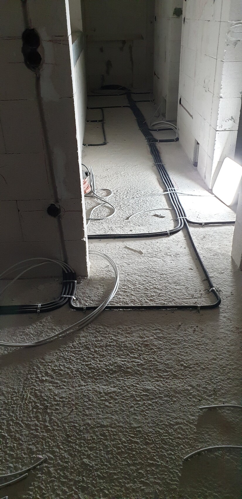 Kabelkanäle und elektrische Leitungen liegen auf dem Betonboden einer Baustelle zwischen Wänden.