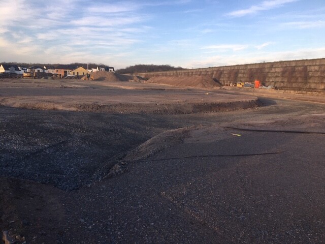 Große Baustelle mit Erd- und Kiesflächen, Häuser am Horizont, offener Himmel.