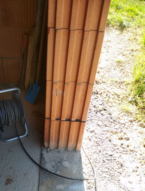 Rote Bauziegel stehen senkrecht an einer Türöffnung; Schlauchrolle und Schaufel daneben.