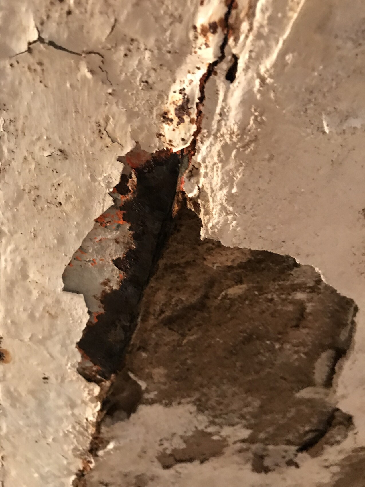 Nahaufnahme einer rissigen Wand mit abblätterndem Putz, Rost und dunklem Spalt.