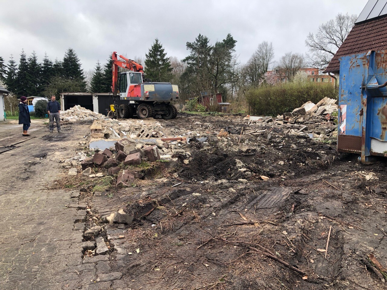 Bau- und Abrissbaustelle mit Bagger, Geröll, blauer Container rechts, zwei Personen links