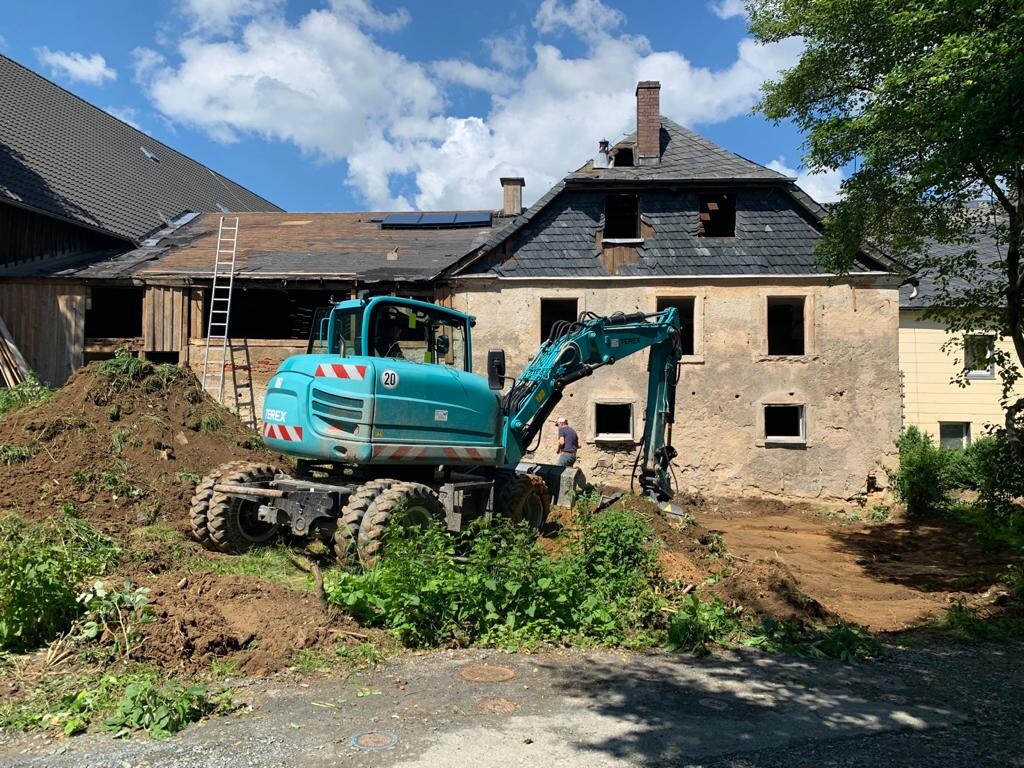 Türkisfarbener Bagger bei Abriss eines alten Hauses, Erdhaufen und Arbeiter sichtbar.