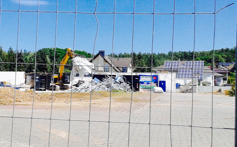 Baustelle mit Bagger und Abrissmüll hinter Zaun, Wohnhäuser im Hintergrund.