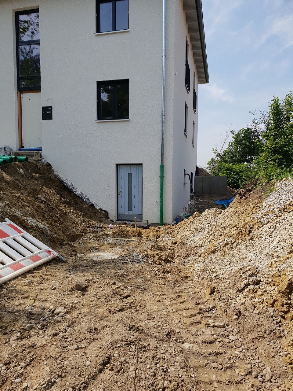 Neubauhaus von außen; Baugrube, Erdaushub, Schutt und grüne Rohre am Fundament.