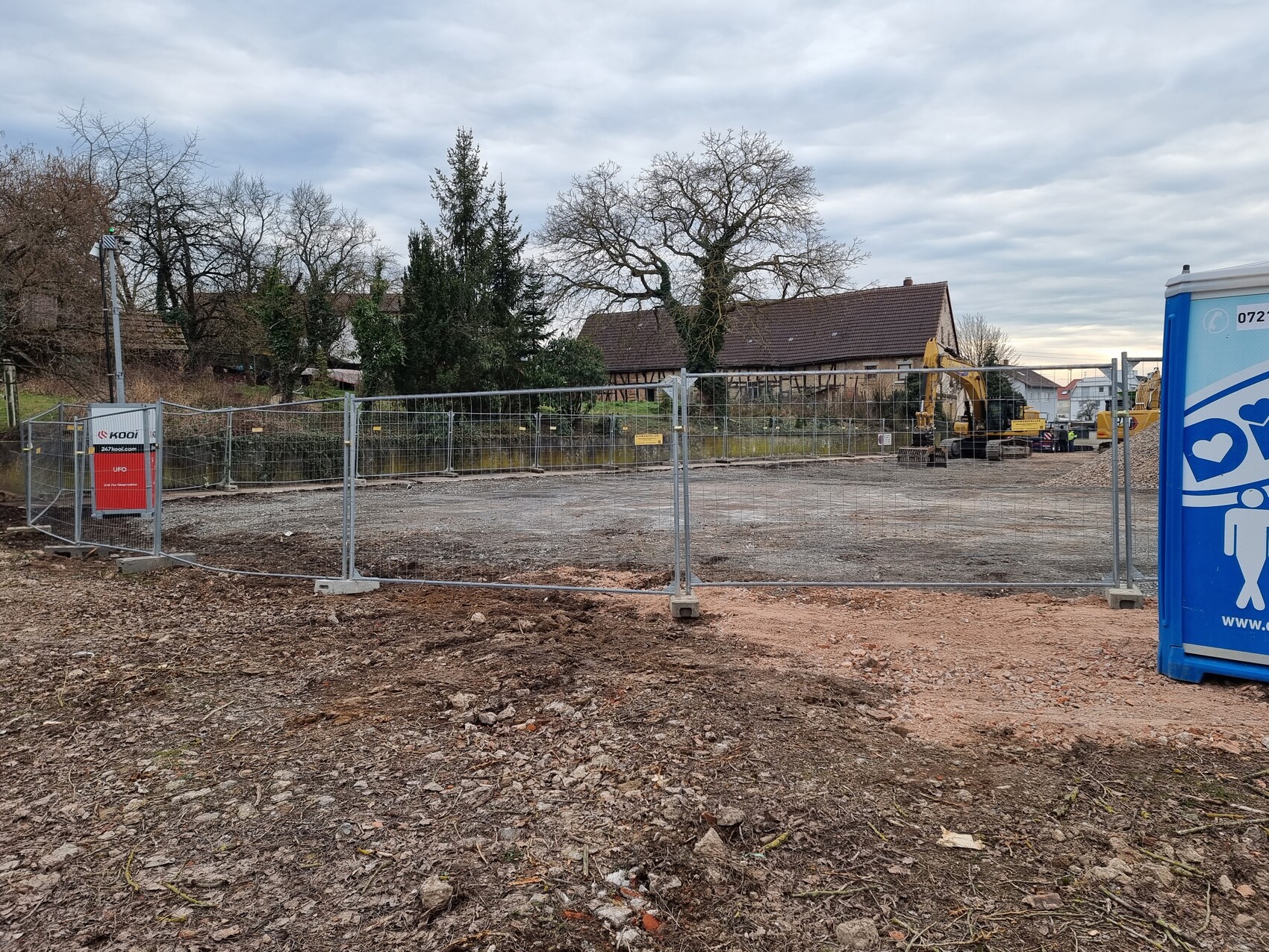 Baustelle hinter Metallzaun mit Bagger, Baucontainer und leerem Erd- bzw. Kiesgelände unter Wolken.
