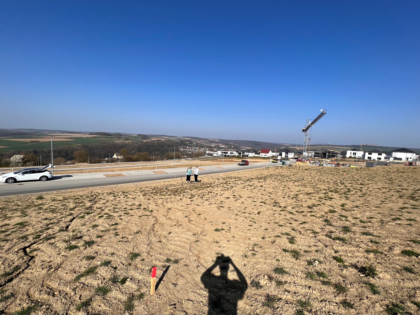 Baugebiet mit Kran, Bauarbeiten, weiße Häuser, Auto am Straßenrand, offene Landschaft.