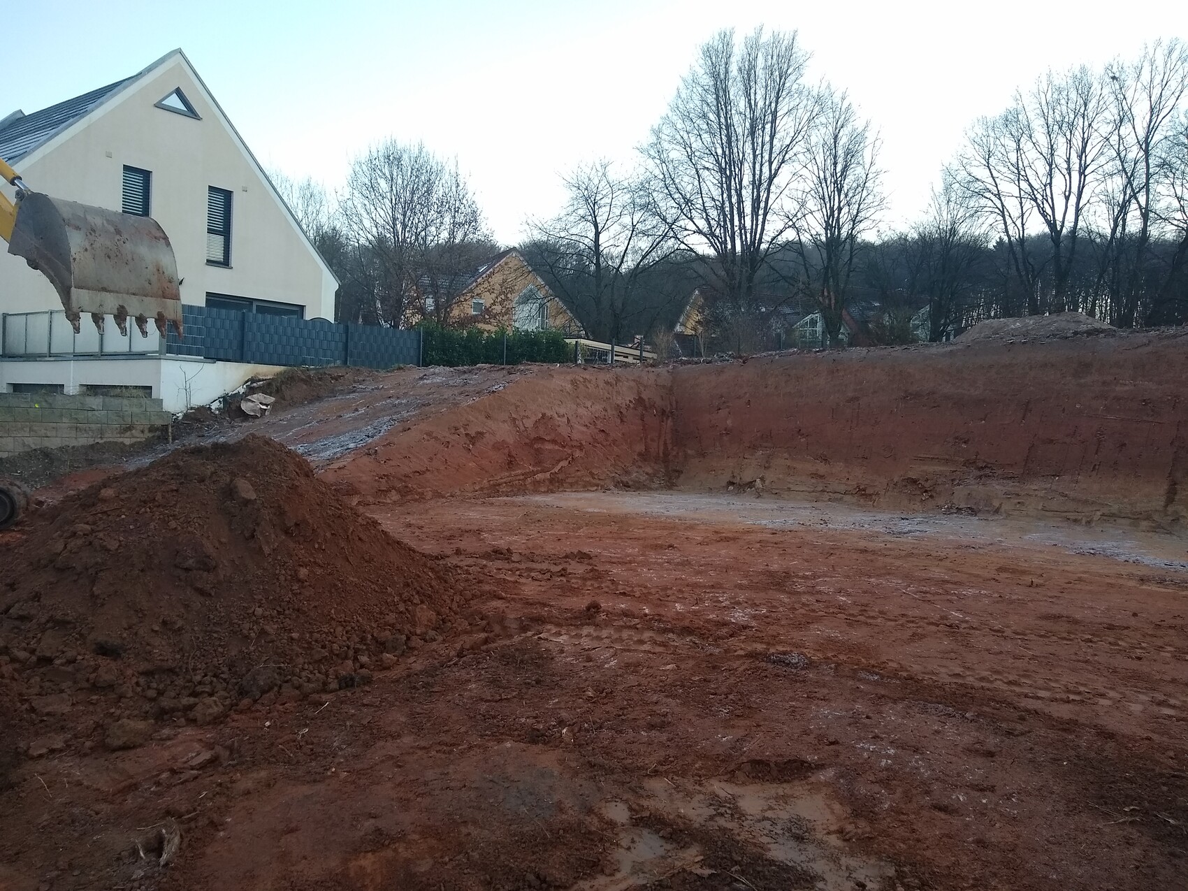 Großer Baugraben mit rotem Erdreich, Schaufel links, Häuser und Bäume im Hintergrund