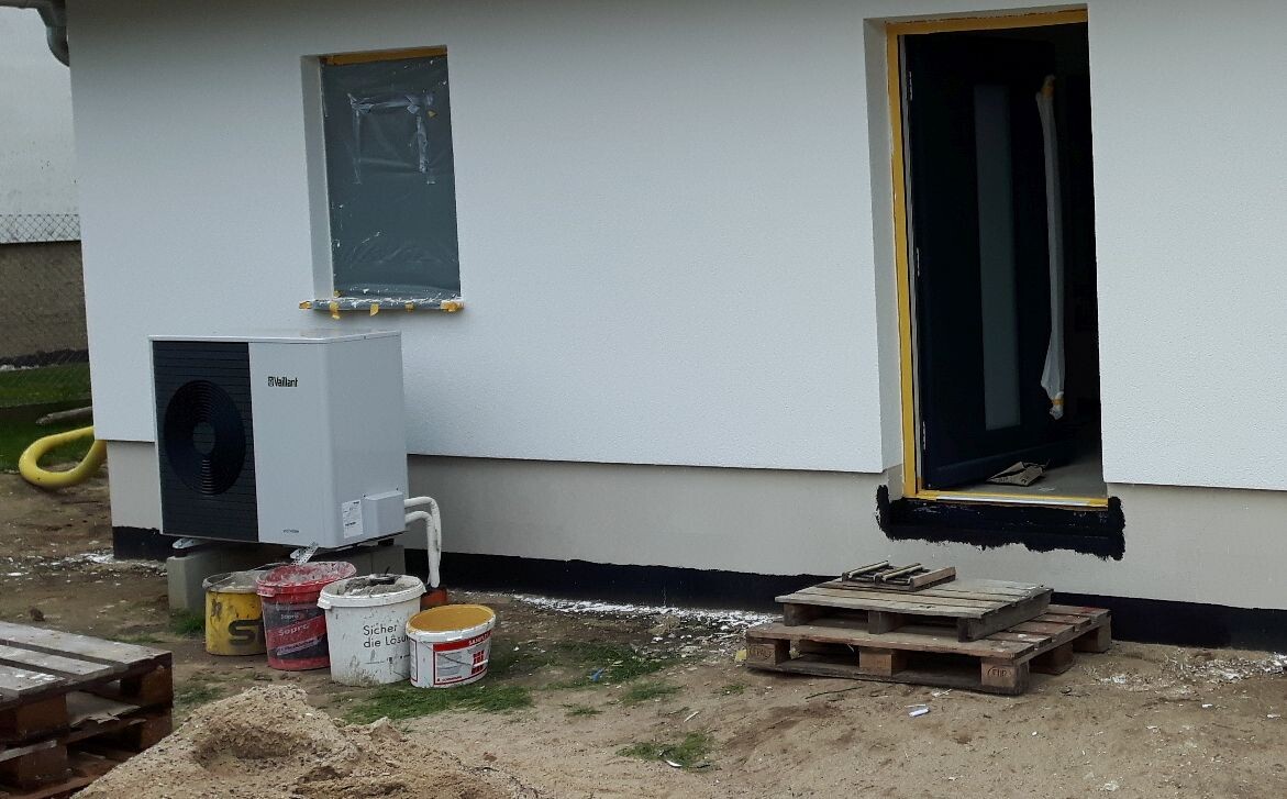 Außenansicht eines Baugebiets: Wärmepumpen-Außengerät, Eimer, Paletten, offener Eingang.
