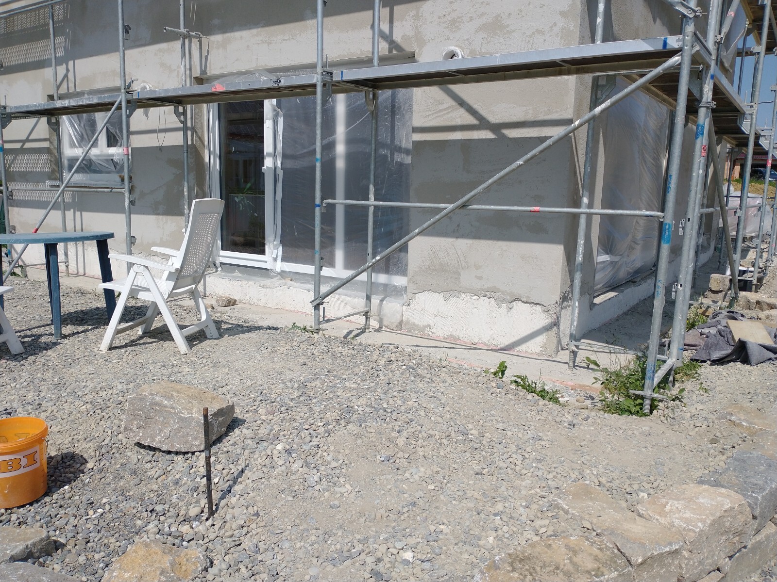 Außenansicht einer Baustelle: Gerüst, verpackte Fenster, Kiesboden, weißer Gartenstuhl.