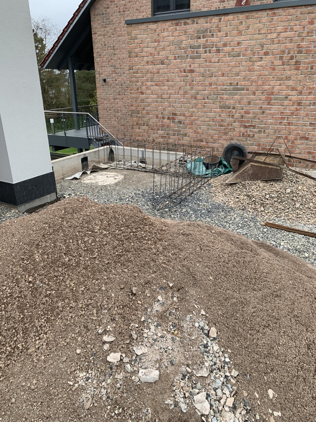 Baustelle vor rotem Ziegelhaus: Erd- und Kieshaufen, Bewehrung aus Stahl, Schubkarren.