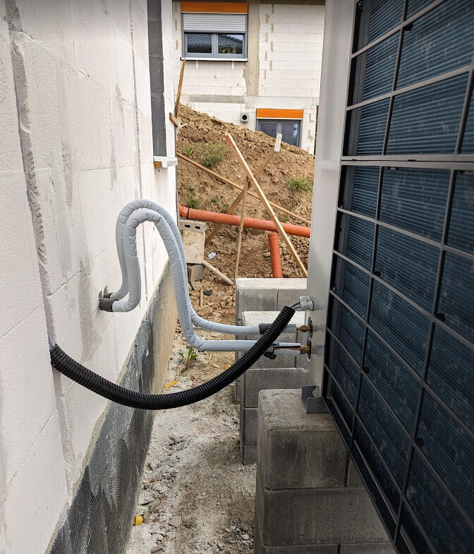 Außenansicht einer Baustelle: Rohre und Kabel verlaufen an einer Wand; Erdarbeiten im Hintergrund.