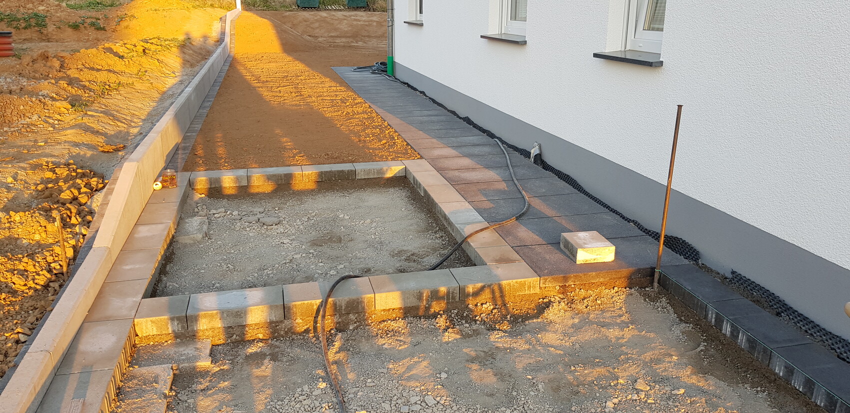 Baustelle am weißen Haus: Pflastersteine werden verlegt, Graben, Kabelschlauch, Schatten.