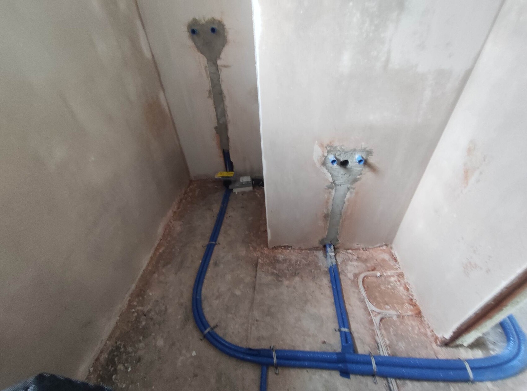 Unbearbeiteter Bauraum: blaue Wasserleitungen verlegt, zwei Wanddurchbrüche mit Rohren sichtbar.
