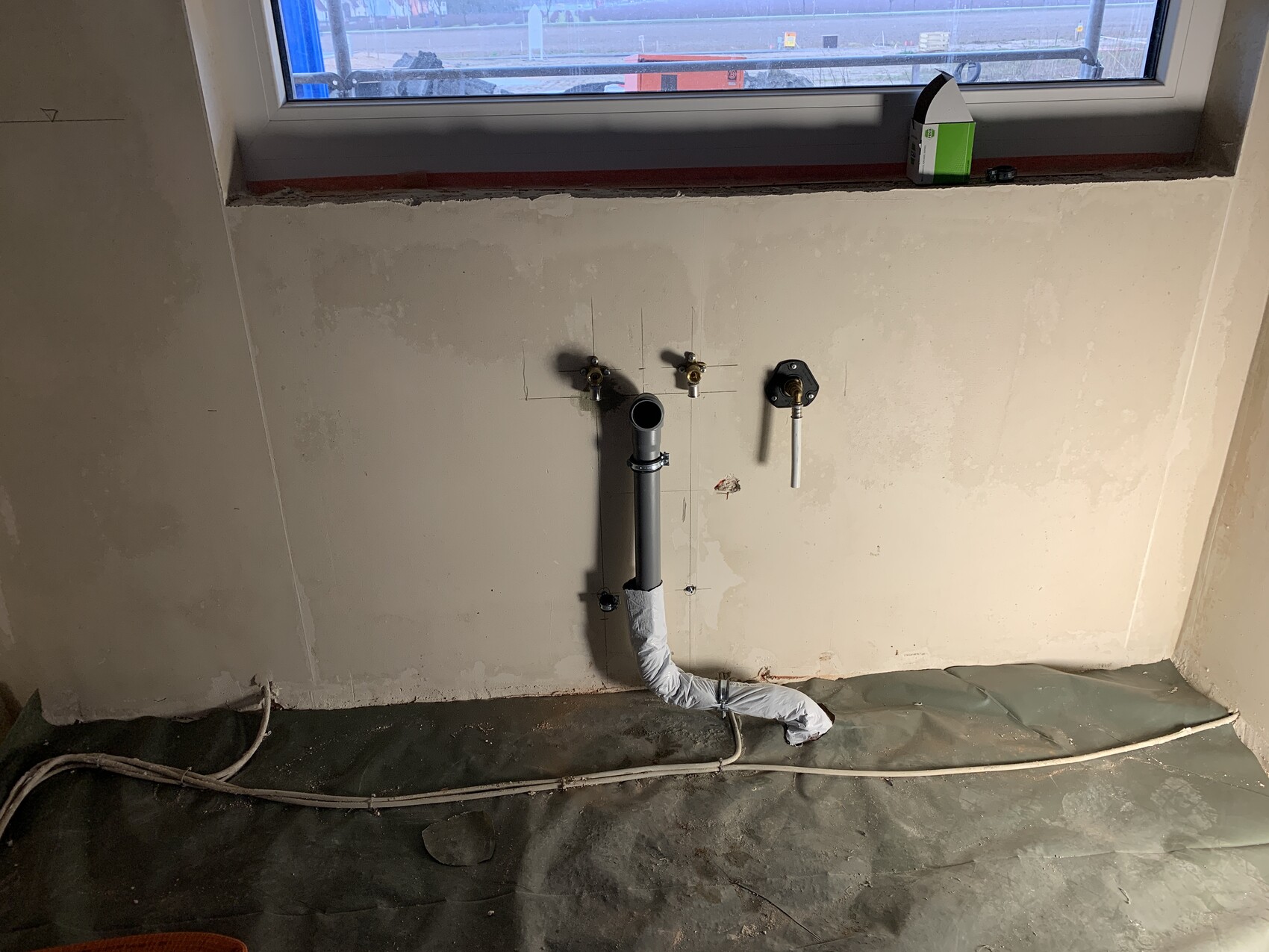 Freigelegte Sanitärinstallation an Wand: Abflussrohr, zwei Ventile, isolierte Leitung und Fenster oben.