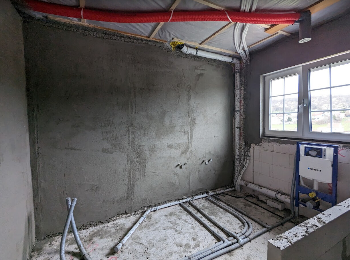 Rohbau eines Badezimmers mit sichtbaren Rohren, Putzwand und Fensterblick.