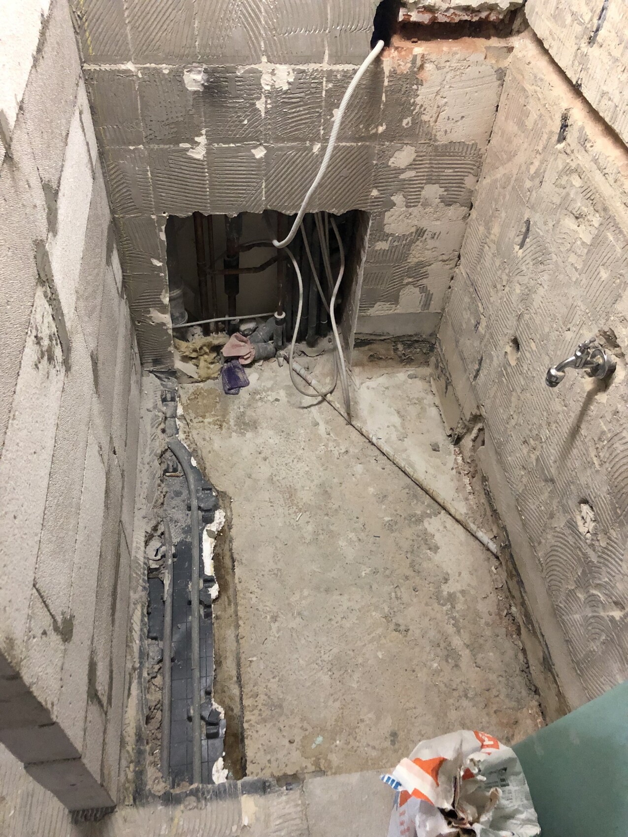 Kleiner Badezimmerrohbau mit offenen Sanitaerrohren, Kabeln und rauen Wänden.