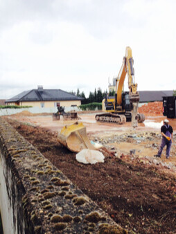 Baustelle mit Bagger beim Fundamentausheben; Erdaushub und Steinmauer am Rand.
