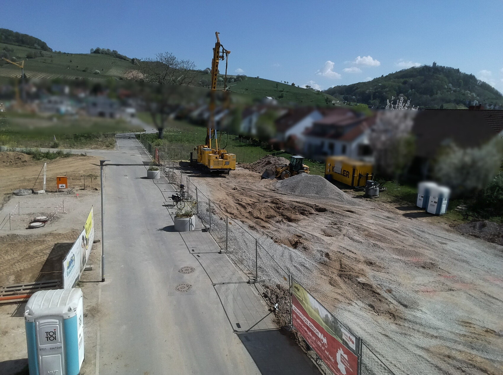 Baustelle mit gelbem Kran, Bagger und Kieshaufen neben Straße in ländlicher Umgebung.