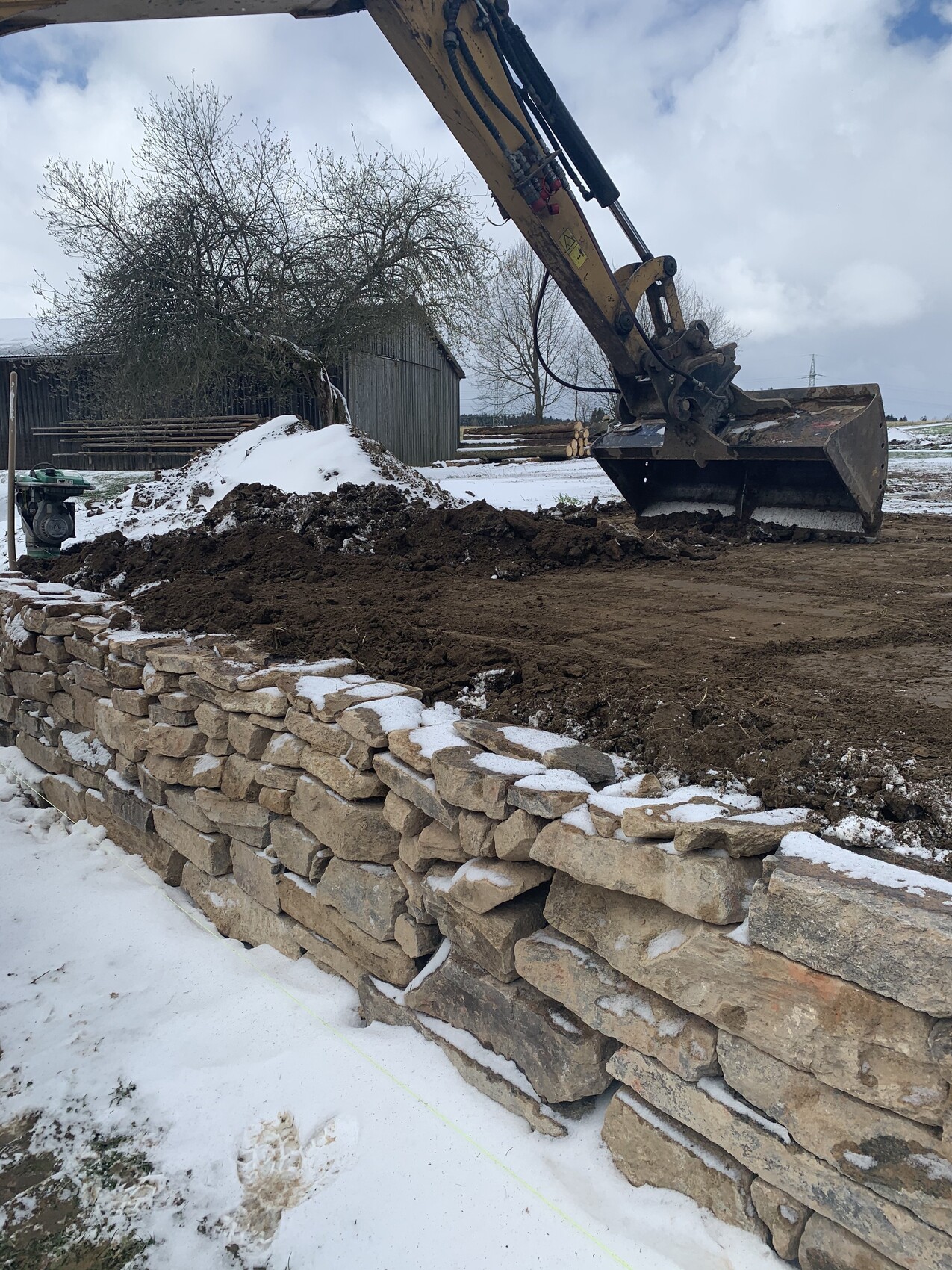 Große Baumaschine mit Schaufel hebt Erde über eine Natursteinmauer; Schnee liegt am Boden.