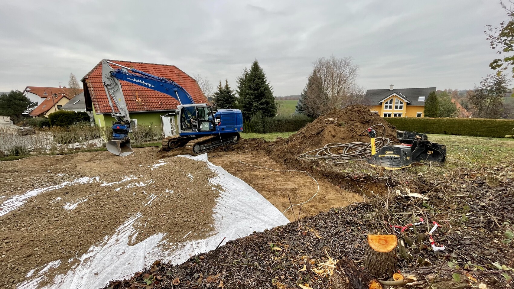 Blaue Bagger-Arbeitsmaschine gräbt eine Baugrube; Erdmassen, Kabelrollen, Wohnhäuser im Hintergrund.
