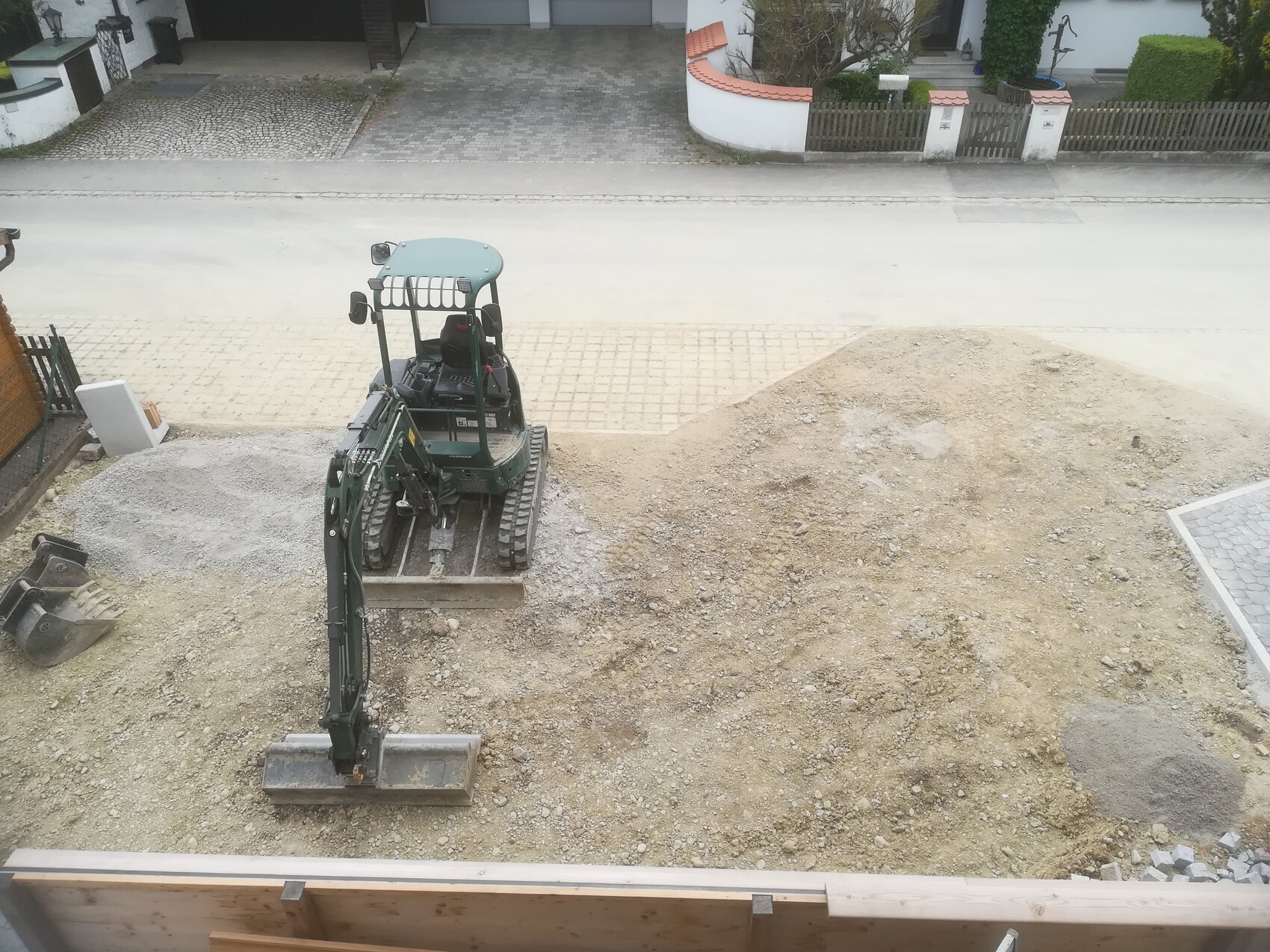Kleiner Bagger auf einer Baustelle neben Wohnhäusern, großer Erd- und Kieshaufen.