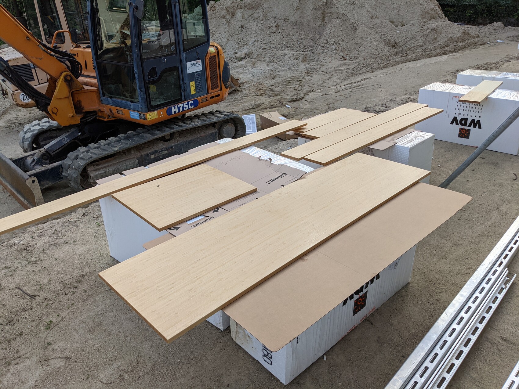 Kleiner Bagger auf Baustelle; Holzbretter liegen auf weißen Styroboxen neben Metallstangen.
