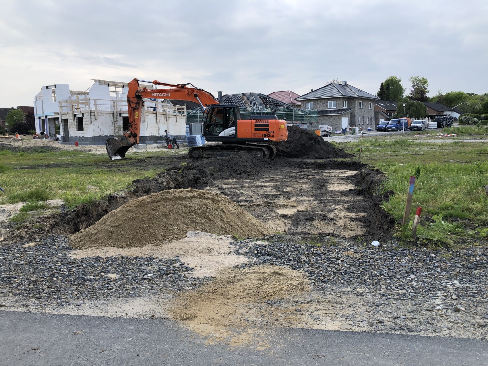 Baustelle mit orangeem Bagger, Graben, Sandhaufen; Wohnhäuser im Hintergrund.