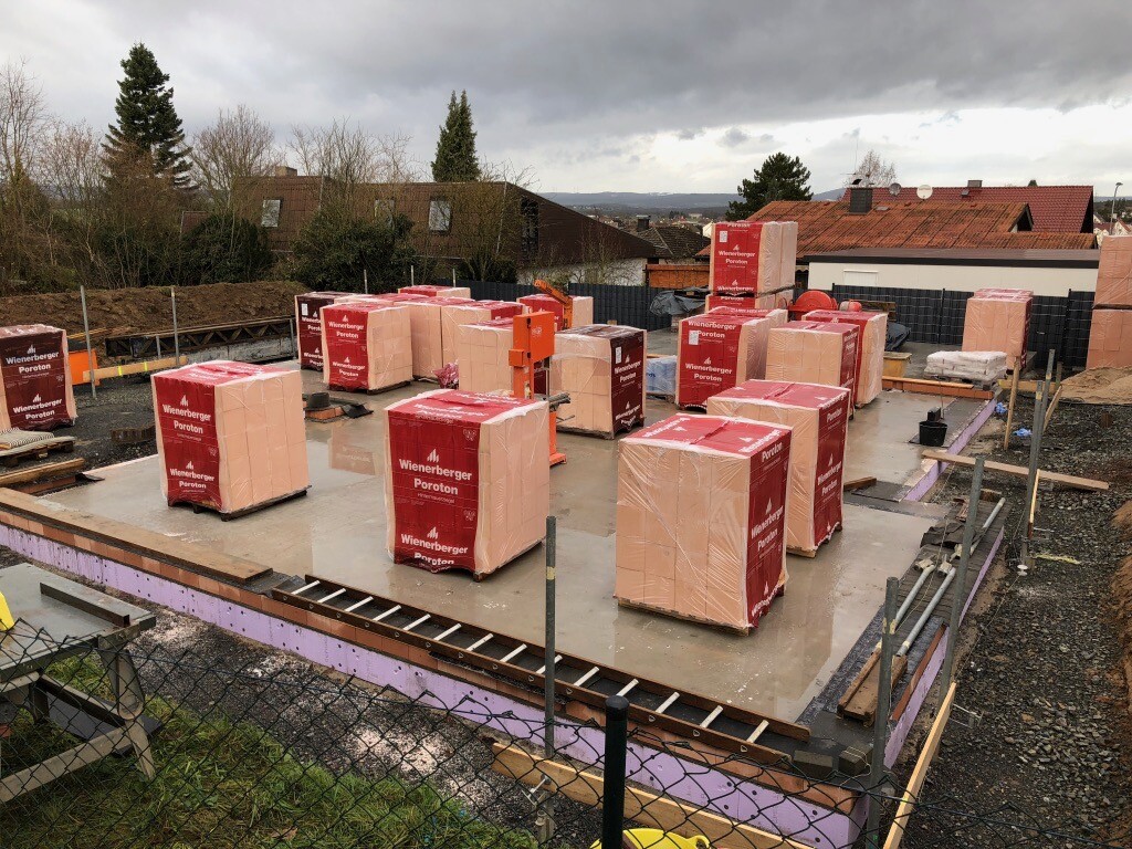 Baustelle mit Paletten aus Wienerberger Poroton-Ziegeln auf der Betonbodenplatte, umzäunt.