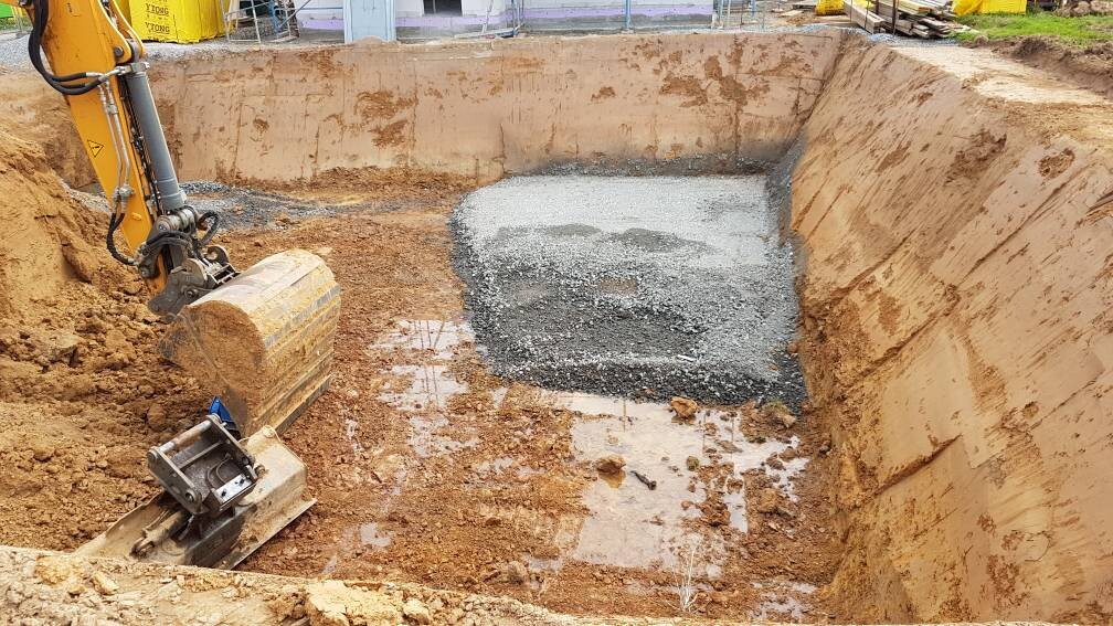 Aushubgrube auf Baustelle; gelber Baggerarm mit Schaufel neben Kiesbett.