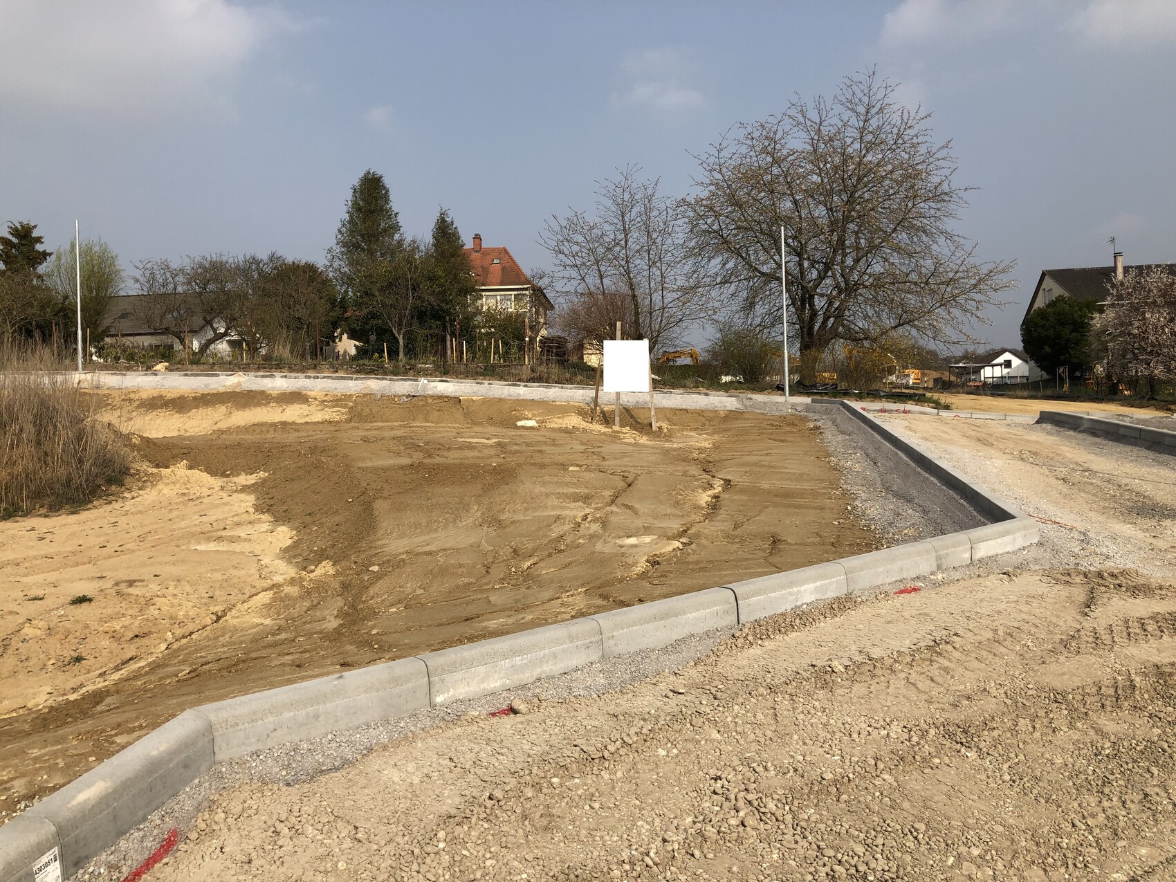 Baustelle mit freiem Erdreich, neuen Bordsteinen, Wohnhäusern und Bäumen im Hintergrund.