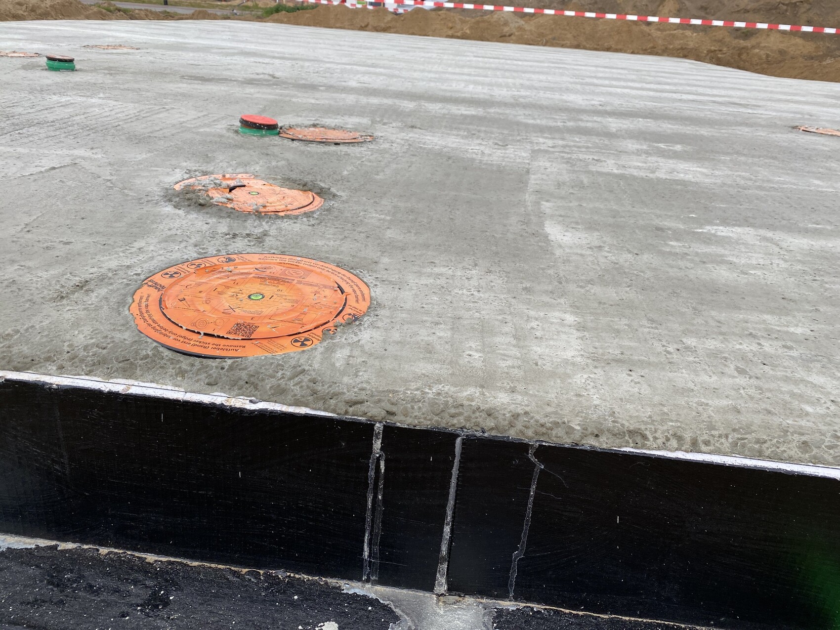 Baustelle mit mehreren orangefarbigen runden Deckeln auf Betonfundament; Absperrband im Hintergrund.