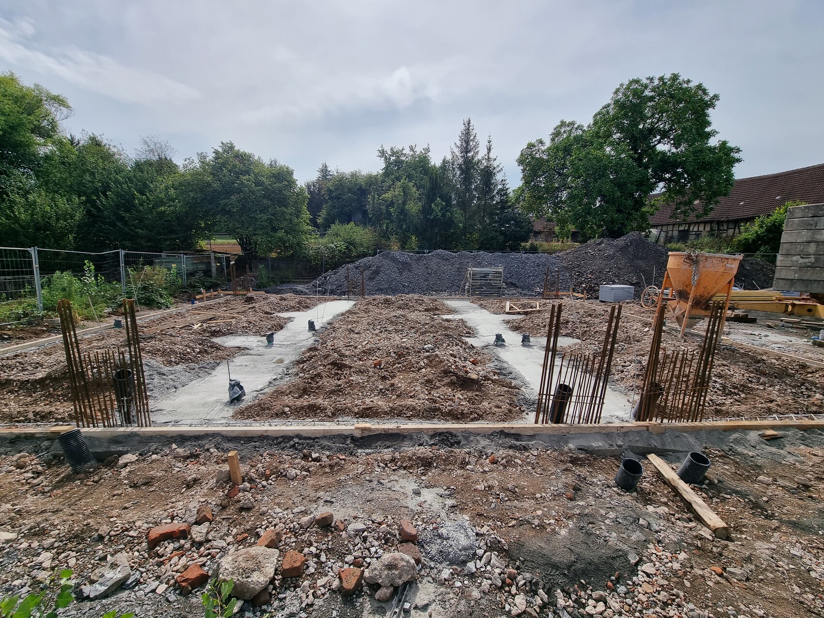 Baustelle mit Fundamentgräben, Bewehrungsstahl ragt hoch, Betonboden, Kies, Baugerät, Bäume.